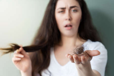 Young stressed woman hold the tips of the brunette hair and shows a bunch of fallen hair. gray background. Concept of alopecia and hair problems.の写真素材