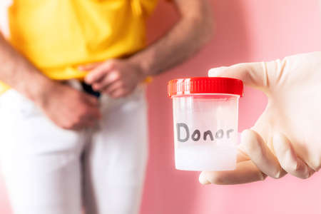 Doctor's hand in a medical glove holds a plastic jar with a sample of sperm. In the defocused background a man shyly zips up his jeans. The concept of sperm donation and artificial insemination.の写真素材