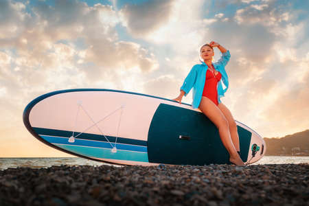 A sexy tanned woman poses sitting at sup board. Sunset in the background. bottom view. copyspace....