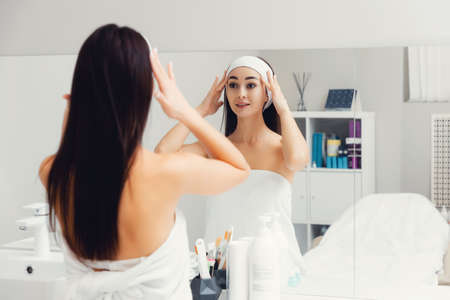 Caucasian young beautiful smiling woman looking at her face in the mirror. back view. The concept of skin care and rejuvenation.の写真素材