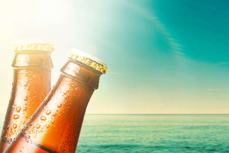 Close-up of the necks of two wet glass bottles of dark beer against the background of the ocean and the sky. copyspace. Alcoholic beverages on vacation.の写真素材