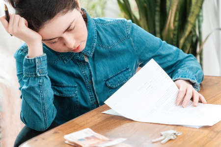 Stressed young woman is sitting at a table with documents, money and keys. The concept of problems with mortgages and loans.の写真素材
