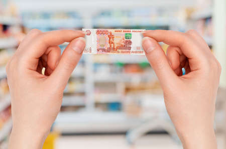 Close up of female hands holds a small five-thousand rubles banknote with her fingers. Blurred showcase of store in the background. The concept of economic crisis and inflation.の写真素材