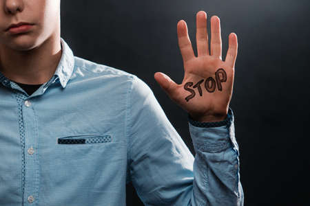 The concept of domestic violence. A sad teenage boy shows a palm up that says STOP. close up. Black background.の写真素材