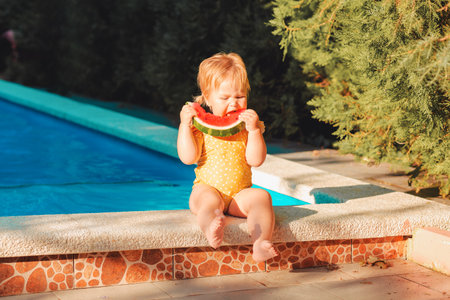 Funny caucasian baby girl in a yellow swimsuit eats a slice of watermelon with an appetite, sitting by the pool. summertime concept.の写真素材