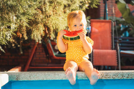 Cute Caucasian baby girl in a yellow swimsuit eats a slice of watermelon while sitting by the pool....