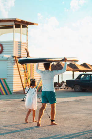 summer family activity. Father and daughter are walking along the beach. Man holding a sup board. back view. sunset light. vertical.の写真素材