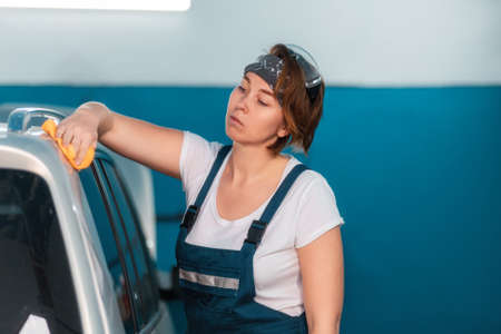 Portrait of a caucasian young woman in a blue coveralls wiping a car with a rag. Car and workshop in the background. Work in an auto repair shop and garage.の写真素材