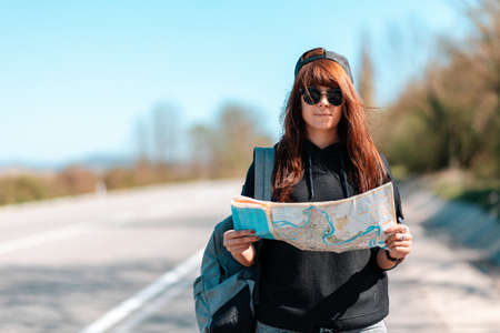 Local Travel and hitchhiking. Portrait of a young woman in a cap and sunglasses, holding a paper map. In the background, the road is blurred. copyspace.の写真素材