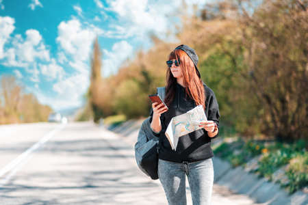 local travel. Young pretty woman in cap and sunglasses stand along the road with a paper map and...