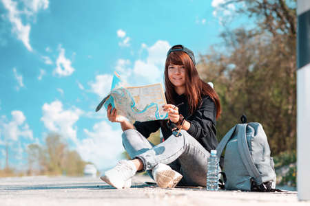 Hitchhiking. A happy hipster girl in cap sitting along the road and holding a paper map. copyspace. Local travel concept.の写真素材