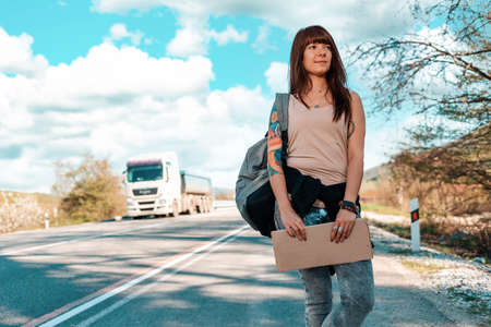 Smiling young woman with tattoed hand holding a cardboard sign. Mock up. car on the background. The concept of local traveling and hitchhiking.の写真素材