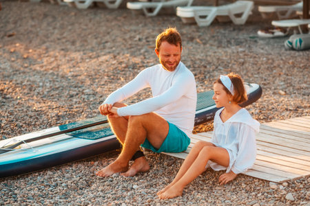 summertime. Father and him daughter sitting and relaxing at the beach. Sup board lie near man. Family vacation at the sea.の写真素材
