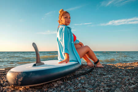 A plump beautiful Caucasian woman poses sitting on a sup board. back view. In the background, the sky is the ocean. The concept of sports recreation by the sea.の写真素材