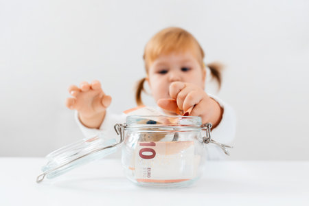 Little cute girl is sitting at a table with a jar filled with money and pulls cash from in it. close up. The concept of finance education and investment.の写真素材