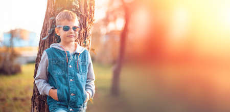 A lonely boy with glasses is standing near a tree. There is a park in the background. copyspace. web...