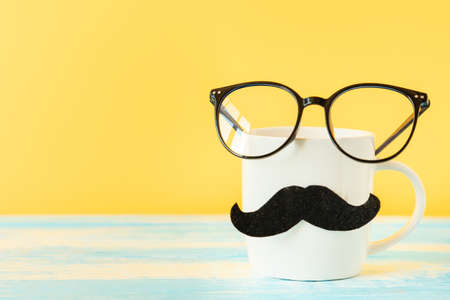 Close up of white ceramic cup with a felt mustache and eyeglasses on yellow background. copyspace. Concept of Father's day and Movember.の写真素材