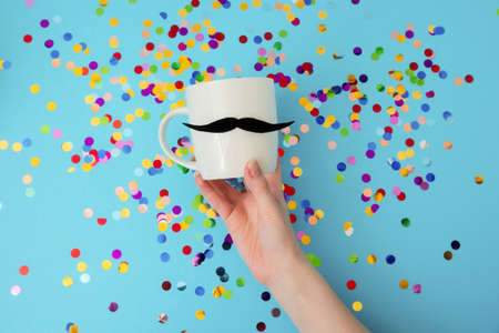 Top view of female hand holding white mug with a felt mustache on a blue background with confetti....