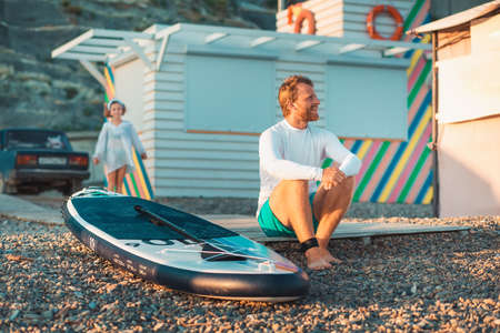 Family vacation at the sea. Father sitting and relaxing at the beach. Sup board lie near man. On the...
