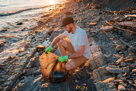 A male volunteer ties a bag full of garbage. In the background-the sea. The concept of environmental conservation and coastal zone cleaning.の写真素材