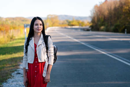 A young beautiful brunette woman in a red dress is standing on the side of the road. In the background, the road goes into the distance and trees. The concept of hitchhikingの写真素材