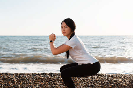 The concept of sport. Young brunette woman doing sports and squats. In the background, the sea and the shore. close up.の写真素材
