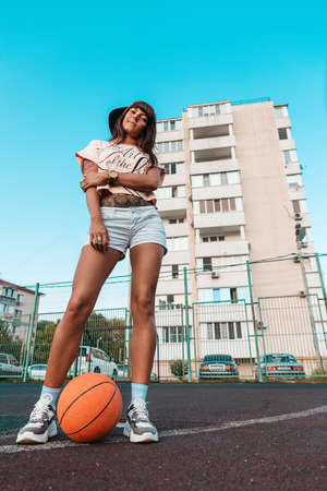A beautiful young woman with tattoos, in a cap, with a basketball in her feet posing for the camera. bottom view.の写真素材