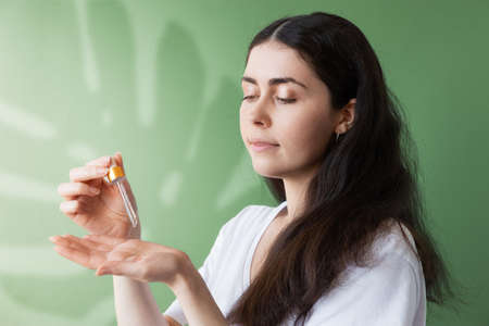 A young beautiful woman applies cosmetic oil from a pipette to the palm of her hand. Green background with a shadow from a monstera leaf. The concept of hair care.の写真素材