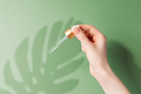 A female's hand holds a pipette with cosmetic oil for skin care. Green background with a shadow from...
