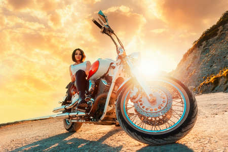 A beautiful confident woman poses sitting on a motorcycle, against the background of an epic sunset....