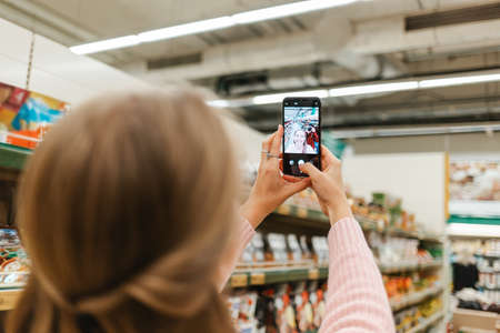 Mobile and social networks. lond woman taking a selfie on smartphone in the grocery store. Rear view from the head.の写真素材