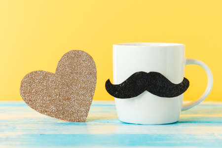 Close up of white ceramic cup with a felt mustache and sparcle golden heart on yellow background. copyspace. Concept of Father's day and Movember.の写真素材