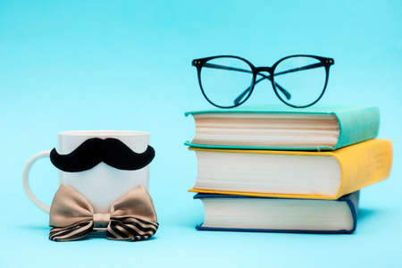 White mug with a felt mustache and silcy bow tie on the blue background with stack of books and glasses. copyspace. Concept of Father's day.の写真素材