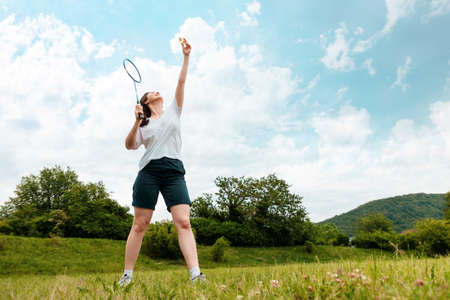 A young sporty woman holds a racket in her hand, preparing to hit a shuttlecock. copyspace. Park on the background. Badminton game and leisure activities.の写真素材