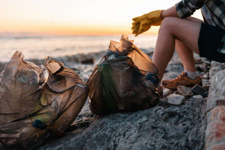 Volunteer sits on the beach with bags full of garbage. In the background, the ocean and sunset. close up. The concept of cleaning the wild beach.の写真素材