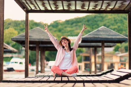 Happy young woman in summer clothes and a straw hat is sitting on a sunbed and shows a victory gesture. Beach umbrellas in the background. The concept of summer vacation.の写真素材
