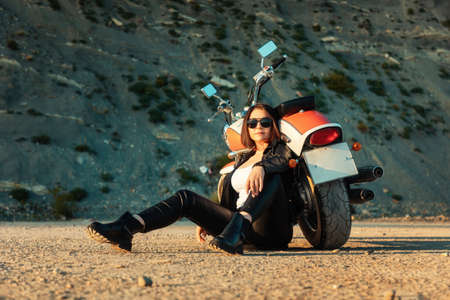 A cool adult woman in a leather motorcycle outfit poses sitting on the ground near a motorcycle. rock in the background. The concept of the Motorcyclist's Day and motorcycle trips.の写真素材