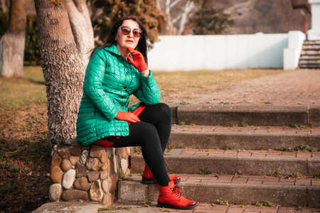 A fashionably dressed senior beautiful Caucasian woman in a green jacket with red accessories is sitting on steps at the park. Active recreation of retired people.の写真素材