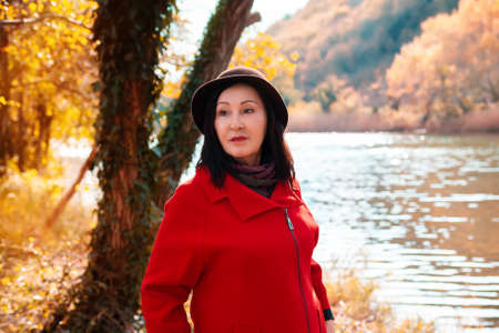 Portrait of elegant beautiful adult woman posing in a hat and red coat. Autumn park and a river in the backgroundの写真素材