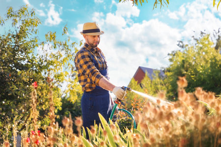 Ecofarm. A bearded, handsome young gardener in uniform and a straw hat, hosing plants and flowers....