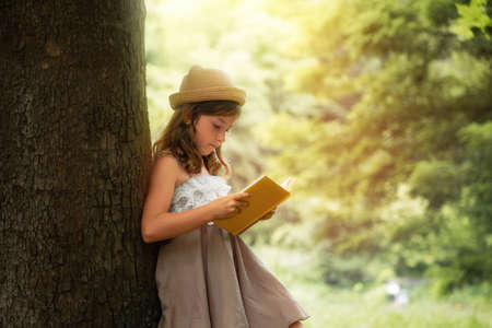 summertime. A beautiful schoolgirl girl in a straw hat and dress is standing by a tree and reading a book. school holidays. The concept of extracurricular children's education.の写真素材