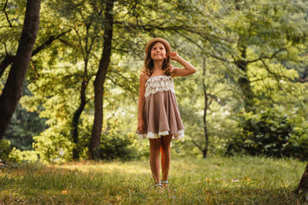 A smiling Caucasian girl in a dress stands on the grass in the park and looks up, holding her hat. Summer holidays. The concept of happy childhood and Children's Day.の写真素材