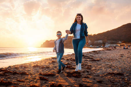 Young smiling mother and her son walking on the beach. In the background is the sea and sunset. The concept of a happy childhood.の写真素材