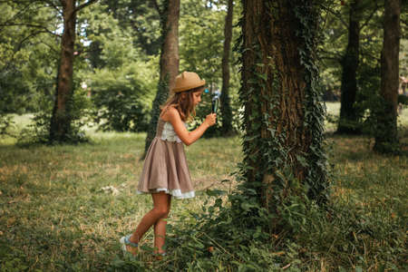 Children's education and curiosity. Caucasian little girl in a straw hat and dress looks at the tree bark through a magnifier. side view. The concept of scouting and childhood.の写真素材