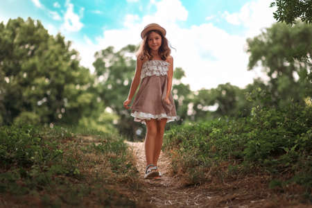 Summer school holidays. Pretty little girl in a straw hat and dress walking in the park. Sky and trees on the background. The concept of childhood and Children's Day.の写真素材