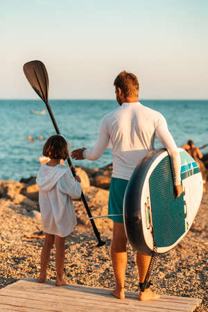 summer holidays. Father and daughter are walking to surf with a sup board. sea in the background. back view. vertical.の写真素材