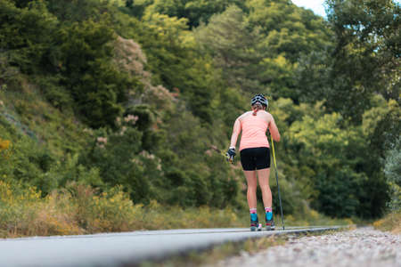 Athletic young woman training on the roller ski at country road, back view. low angle view. Concept of sports competition and biathlon summer workout.の写真素材
