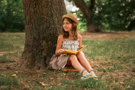 Smiling cute schoolgirl girl in a straw hat and dress is sitting on the grass by a tree holding a book. school holidays. The concept of independent extracurricular children's education.の写真素材