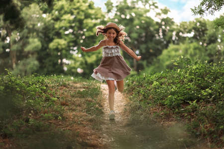 Cheerful schoolgirl in a hat and dress runs from the slope in the park. Summer holidays. The concept of happy childhood and Children's Day.の写真素材