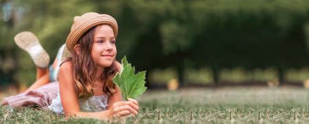 Web banner of ecology and Earth Day with copy space. Portrait of cute smiling girl in a straw hat lies on the lawn and holding a green leaf. The concept of summertime and happy child.の写真素材